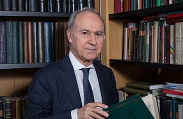 A man in suit with a book in his hand sitting in a library corner with a stack of books in the background