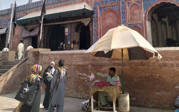Three Muslim women standing outside of the shrine of Pir Shams near a man sitting under an umbrella selling rose petals.