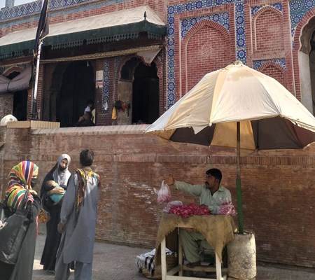 Three Muslim women standing outside of the shrine of Pir Shams near a man sitting under an umbrella selling rose petals.