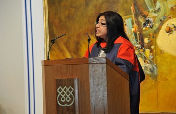 Dr. Laila Halani standing at the podium and delivering a speech in a graduation gown at the social hall of Ismaili Centre, London