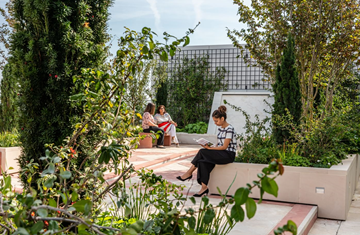 A rear view image of a girl (in the frame) reading and two women (in the blurry background) sitting in the terrace garden of AKC