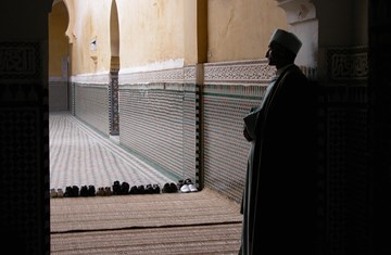 A man dressed in traditional clothes patiently standing in the corner of a prayer hall and praying 