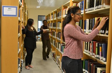 A student picking out a book from the library shelf and a female staff member coordinating with male staff member in the IIS library
