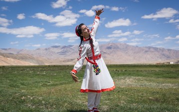 A girl in white Tajikistan dress in a swirling position with eyes closed , standing in a garden with a mountainous scenery background