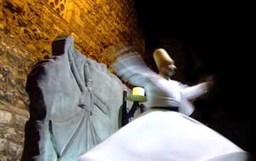 A wall with sculpted dervish carved on wood and a blurry view of a live dervish doing a Sufi dance
