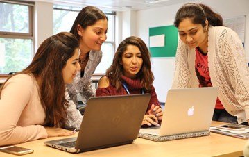 An IIS faculty leaning and observing the work in the laptops with two students sitting and one student standing and listening from the side