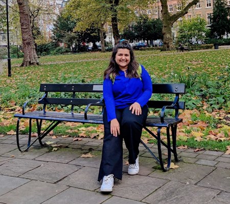 A young lady sat on a bench in a London square with grass and fallen leaves behind her