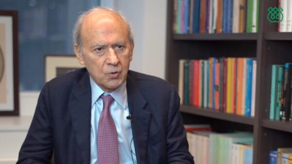 Dr. Farhad Daftary in a black suit talking in a room with a stack of books in the background on the right hand side