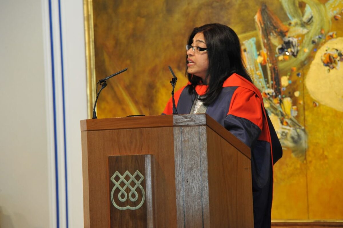 Dr. Laila Halani standing at the podium and delivering a speech in a graduation gown at the social hall of Ismaili Centre, London