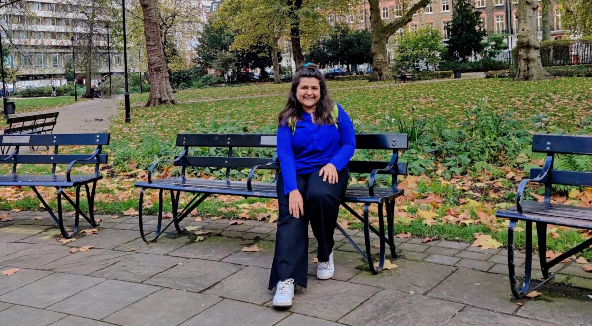 A young lady sat on a bench in a London square with grass and fallen leaves behind her