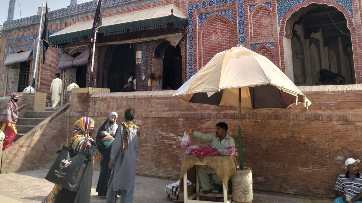 Three Muslim women standing outside of the shrine of Pir Shams near a man sitting under an umbrella selling rose petals.