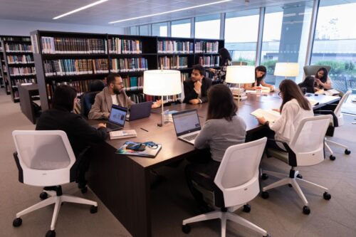 Group of students sitting around a table in library setting with book shelves behind them