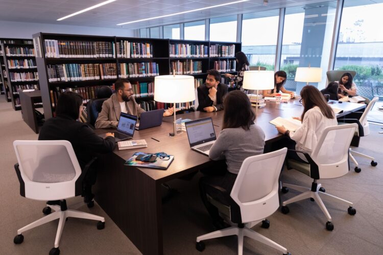 Group of students sitting around a table in library setting with book shelves behind them
