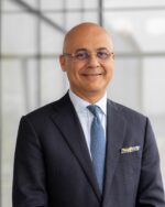 Amyn Kassim Lakha, IIS Governor, smiling in rimless glasses, a navy suit and a patterned pocket square.