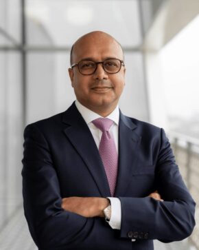 Arif Jamal, IIS Governor, smiling with arms crossed, wearing glasses, a navy suit and a patterned pink tie.