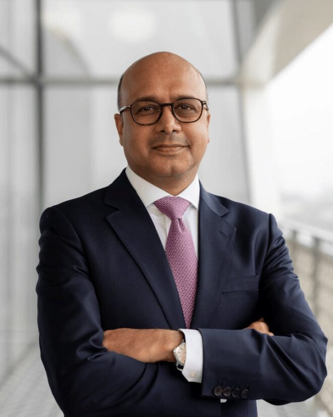 Arif Jamal, IIS Governor, smiling with arms crossed, wearing glasses, a navy suit and a patterned pink tie.