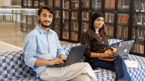 GPISH students Ahmed Faraz and Anila Goulamhussen sit with laptops on patterned seating in front of glass-fronted bookcases.