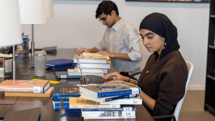 GPISH student Anila Goulamhussen works on a laptop at a library desk stacked with books; a second student reads behind.
