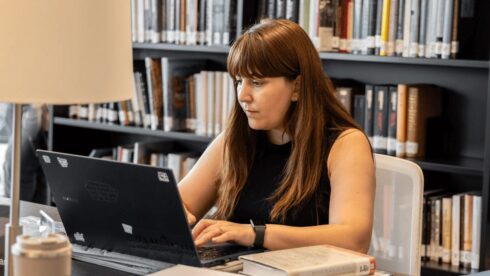 GPISH student Shenida Sultana types on a laptop at a library desk, with shelves of books behind her.