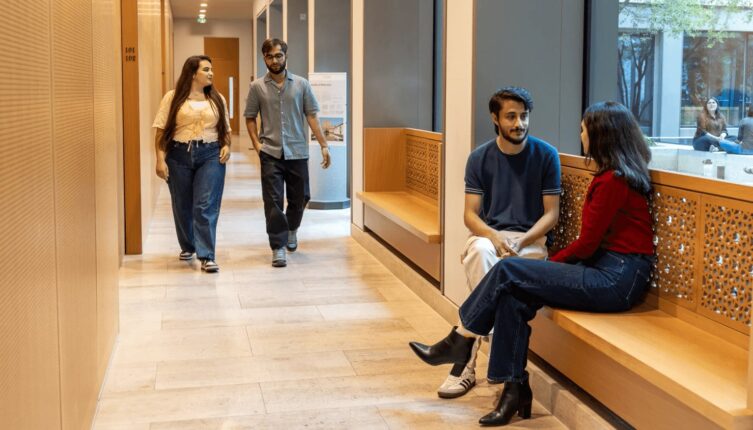 Two GPISH students walk along a corridor at Victoria Hall while two others sit talking on a wooden bench.