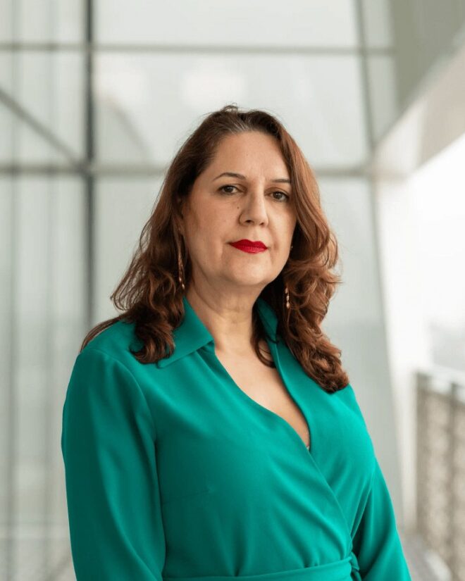Nacim Pak-Shiraz, IIS Governor, with wavy auburn hair, wearing a teal wrap blouse.