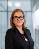 Selina Kassam Ramji smiling, wearing black glasses and a black jacket, in a bright glass-walled interior.