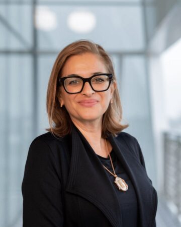 Selina Kassam Ramji smiling, wearing black glasses and a black jacket, in a bright glass-walled interior.