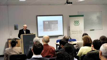 A professor giving a lecture in a room full of people with a projector screen behind him