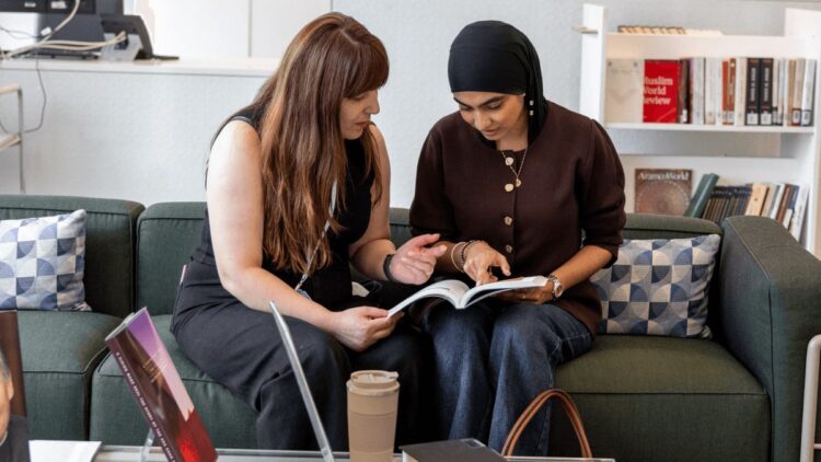 Two GPISH students sit on a green sofa, pointing at a page in an open book they are reading together.