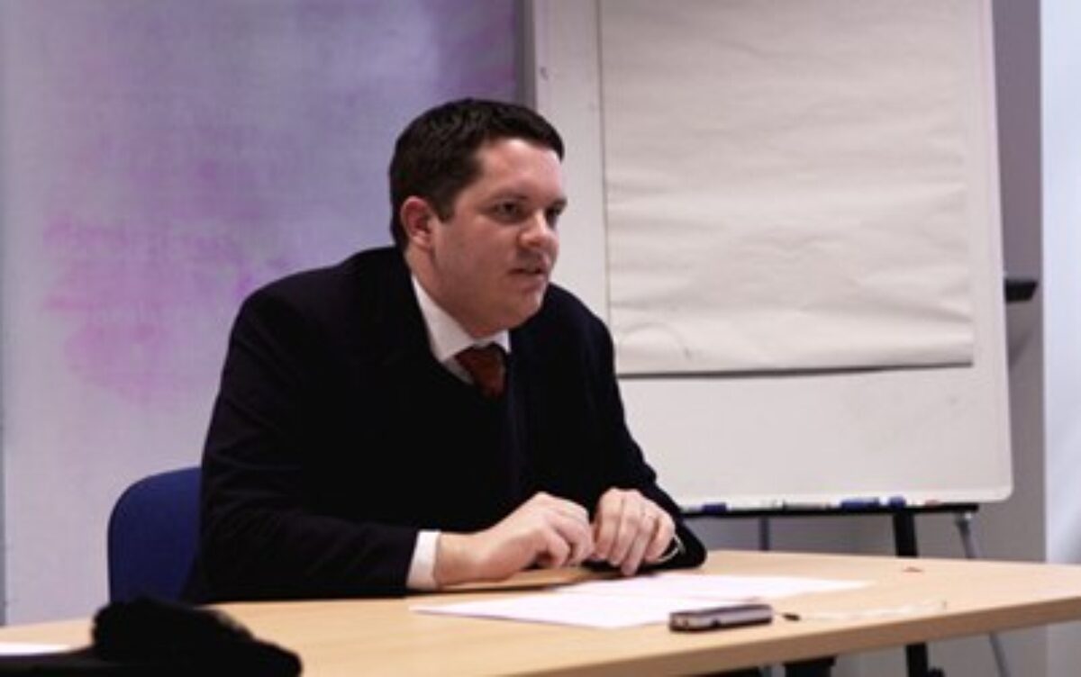 A man in black suit sitting and addressing a audience
