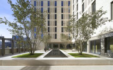 Courtyard with a long reflecting pool, trees and two people seated, framed by a tall patterned brick building.