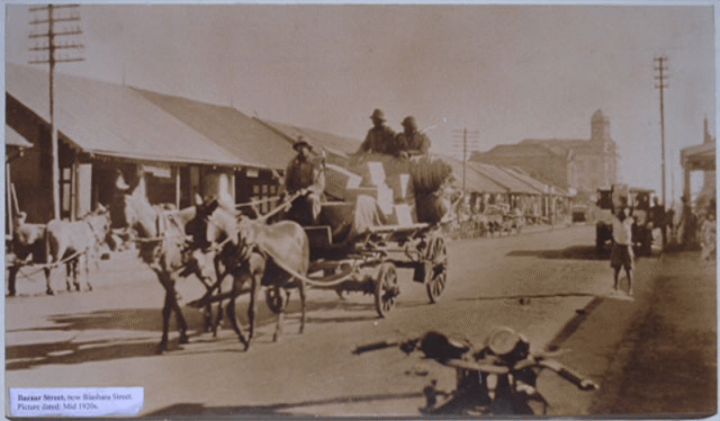 Single-storey businesses, horse-drawn carriages, a motorcycle, and a jeep foreground Bazaar Street