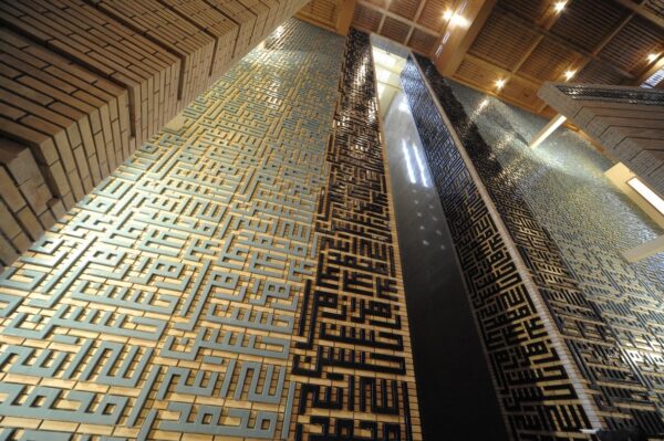 Light, cast across a wall of calligraphic repetition of the names 'Allah', 'Muhammad', 'ʿAli', 'Hasan', 'Husayn', and 'Zahra'
