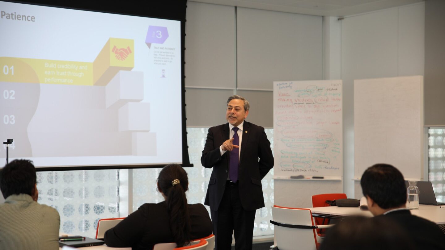 a man in a suit in a classroom, pointing at a projector screen
