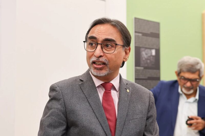 A man in a grey suit with a red tie stands in a formal gallery setting. Informational posters are visible on the wall behind him. Another person in a blue suit stands in the background.