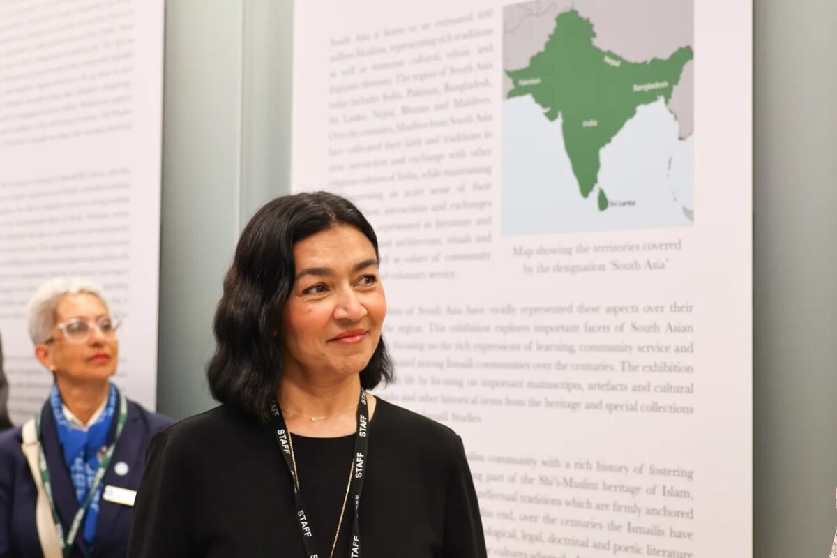 A woman with shoulder-length dark hair wearing a black top and a lanyard stands in front of an exhibit. Another person is visible in the background. The exhibit includes a map of South Asia and text.