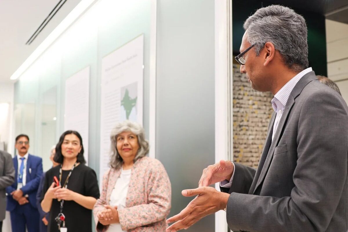 A group of individuals in business attire stand in a modern room with informational displays on the wall. One person is speaking and gesturing with hands while others listen attentively.