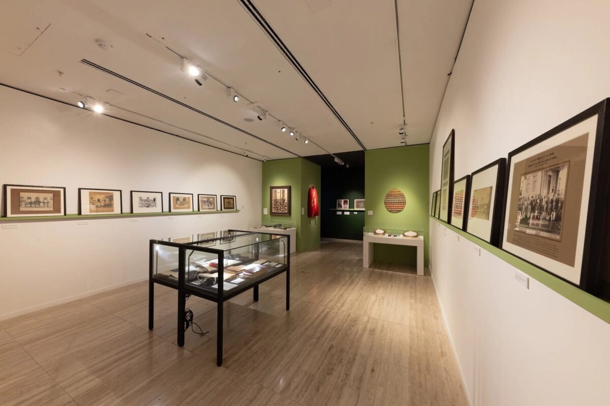 A gallery room with framed pictures on white walls, display cases with artefacts, and a green accent wall. Spotlights illuminate the exhibits.