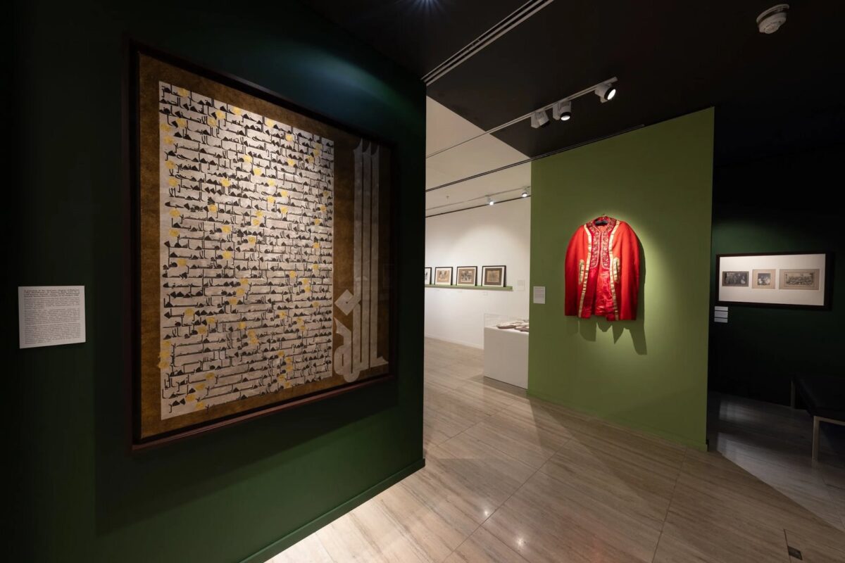 A gallery exhibit showing a framed piece of calligraphy on a green wall and a red garment with gold embroidery. Display cases and framed images are visible in the background.