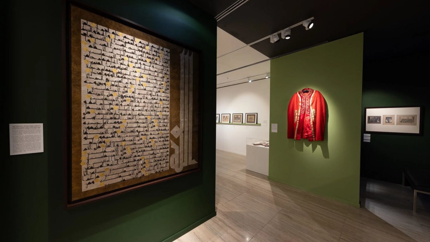 A gallery exhibit showing a framed piece of calligraphy on a green wall and a red garment with gold embroidery. Display cases and framed images are visible in the background.