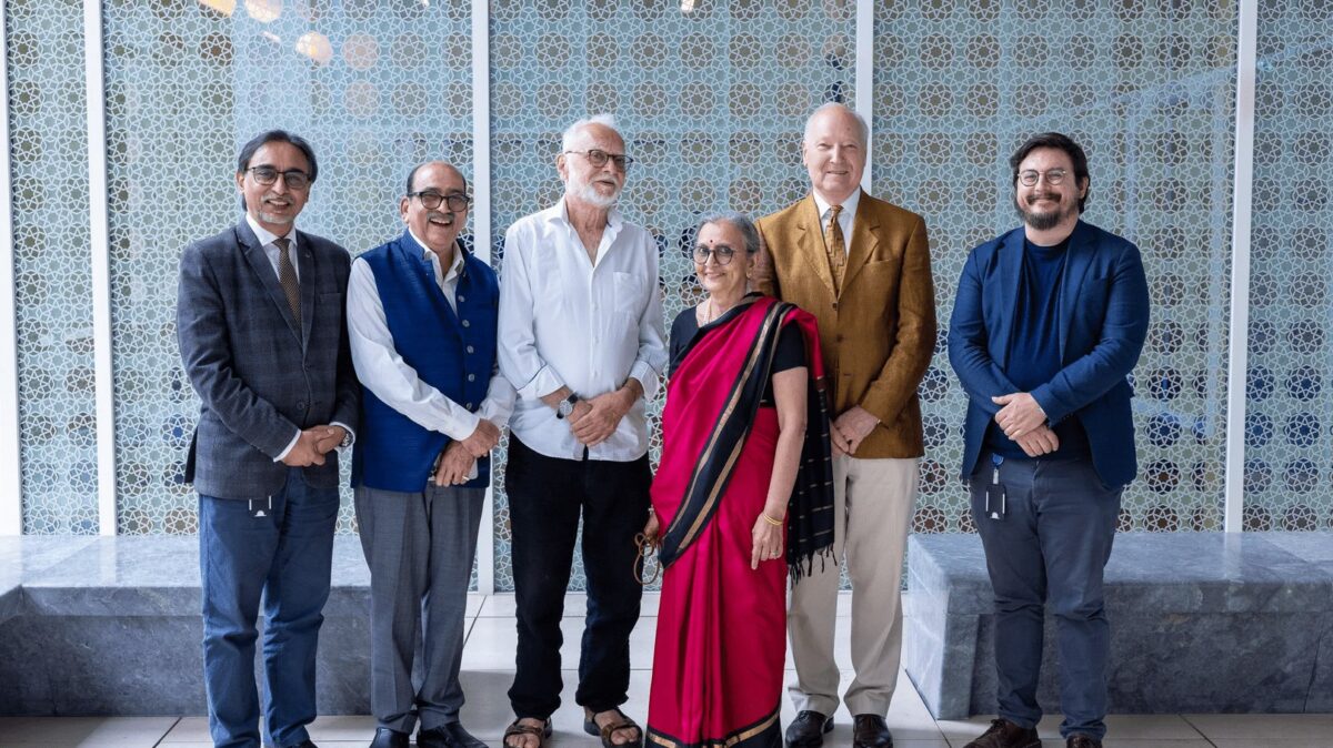 Six people stand in a row facing the camera, dressed in a mix of formal, semi-formal, and traditional attire. The background features a patterned wall with geometric designs.