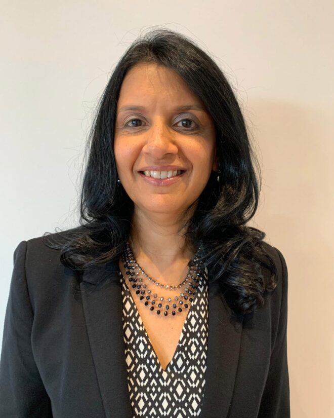 Woman in a black jacked smiling, posing against a plain background. Dr Tasnim Dharamsi