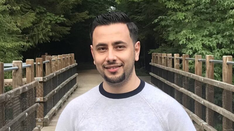 Rami Gargour stands on a wooden footbridge surrounded by trees, photographed outdoors in a natural setting.