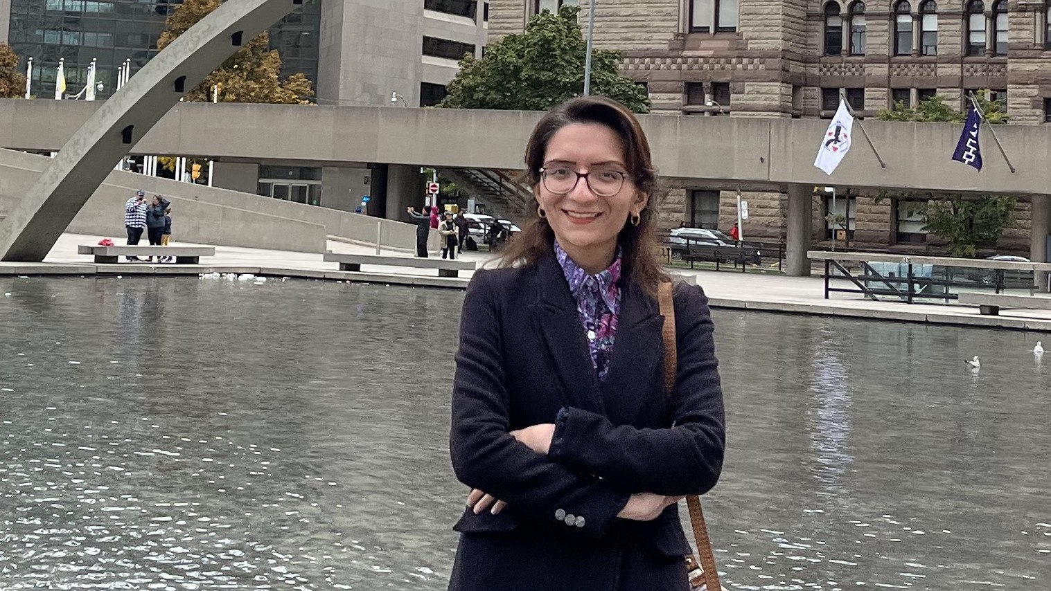 Seddigheh Kardan stands outdoors near a reflective water feature, with urban architecture visible in the background.