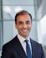 Fayyaz Nurmohamed, IIS Governor, smiling in a navy suit and blue tie against a blurred glass background.