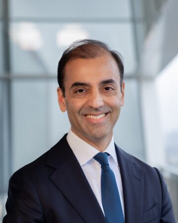 Fayyaz Nurmohamed, IIS Governor, smiling in a navy suit and blue tie against a blurred glass background.