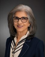 Dr Minaz Fazal with grey hair, in a professional suit, posing against a dark background.