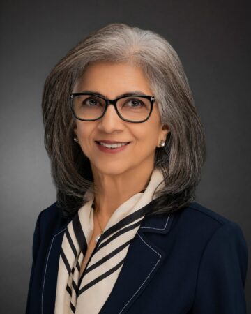 Dr Minaz Fazal with grey hair, in a professional suit, posing against a dark background.