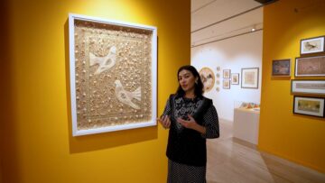 Gallery curator Esen Kaya standing beside a framed artwork of two birds on a yellow wall at the Aga Khan Centre Gallery