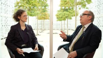 Russell Harris and Tara Woolnough in conversation during a recorded discussion, seated indoors with trees visible through large windows behind them